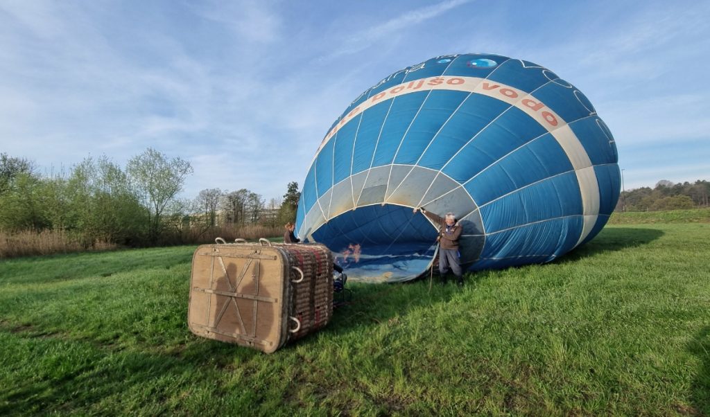 A blue hot air balloon is being inflated on a grassy field beside its wicker basket. Two people stand near the open mouth of the balloon as the burner sends flame inside under a wide pale blue sky.