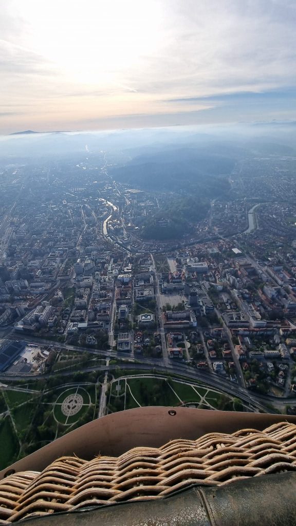 View from a hot air balloon basket over a city at sunrise with roads, buildings, and a winding river fading into blue haze. The woven rim of the basket fills the foreground while the pale sun brightens the sky above distant hills.