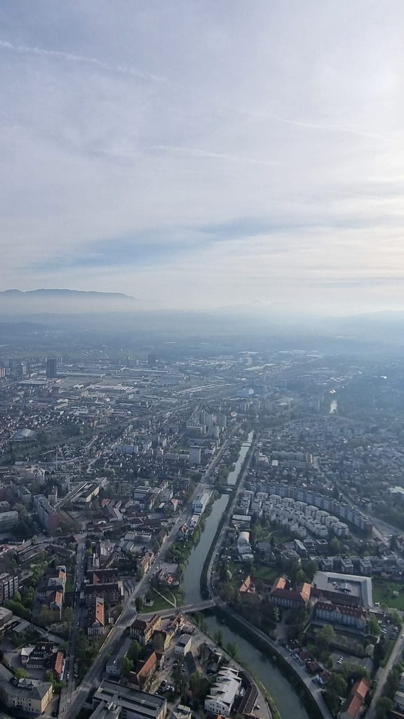 Aerial view of a city with a canal running through dense neighborhoods, apartment buildings, and industrial areas under a hazy morning sky. Low mountains sit along the horizon as the waterway leads into the distance.
