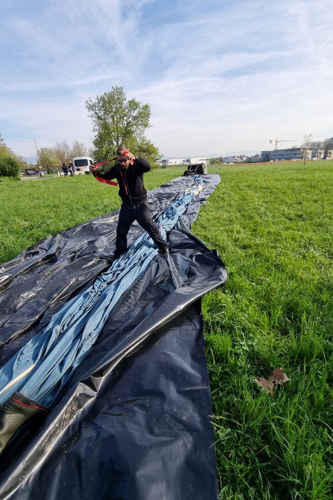 A man stands on a long strip of deflated balloon fabric in a grassy field, pulling a red strap as he helps pack it away. A van, a tree, and low buildings sit in the distance under a streaky blue sky.