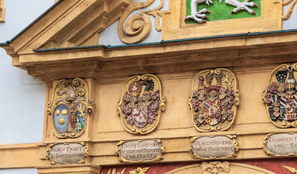 Ornate painted facade detail with carved coats of arms in gold framed panels arranged across a historic building. Decorative name plaques beneath the crests and scrolling trim emphasize the richly preserved old town architecture.