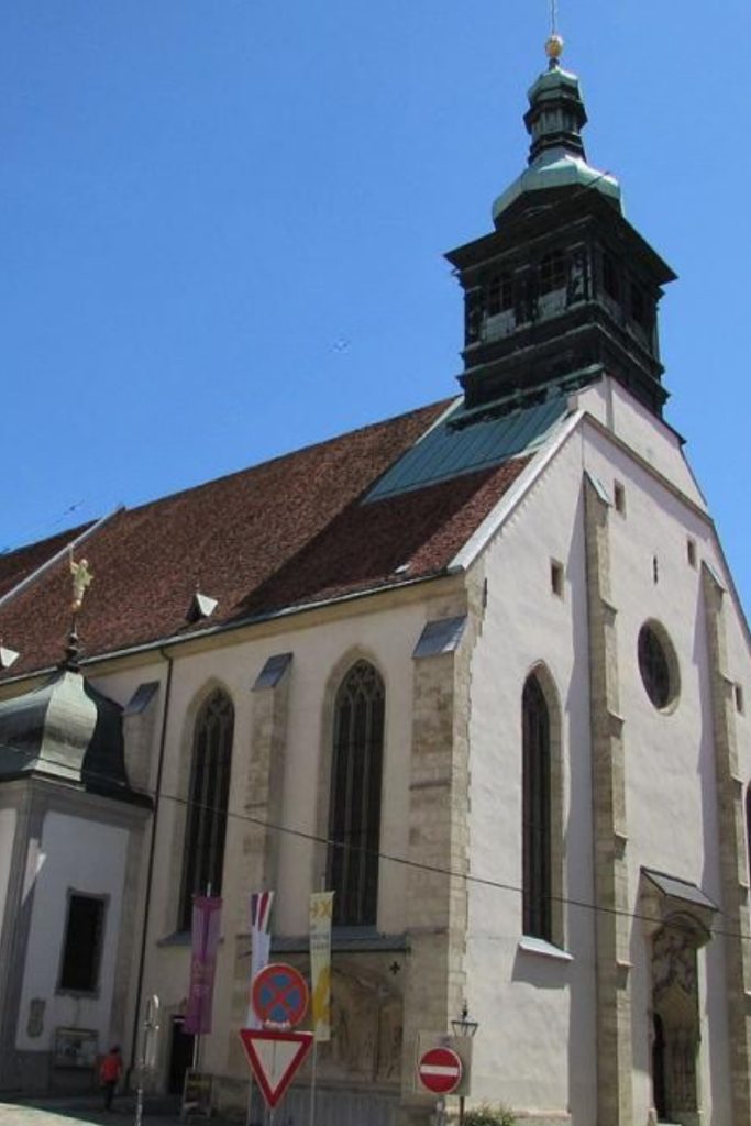 White church with tall narrow Gothic windows and a dark tower rising above a steep red tiled roof under a clear blue sky. Street signs and banners at the base show the church from ground level in the middle of the city.