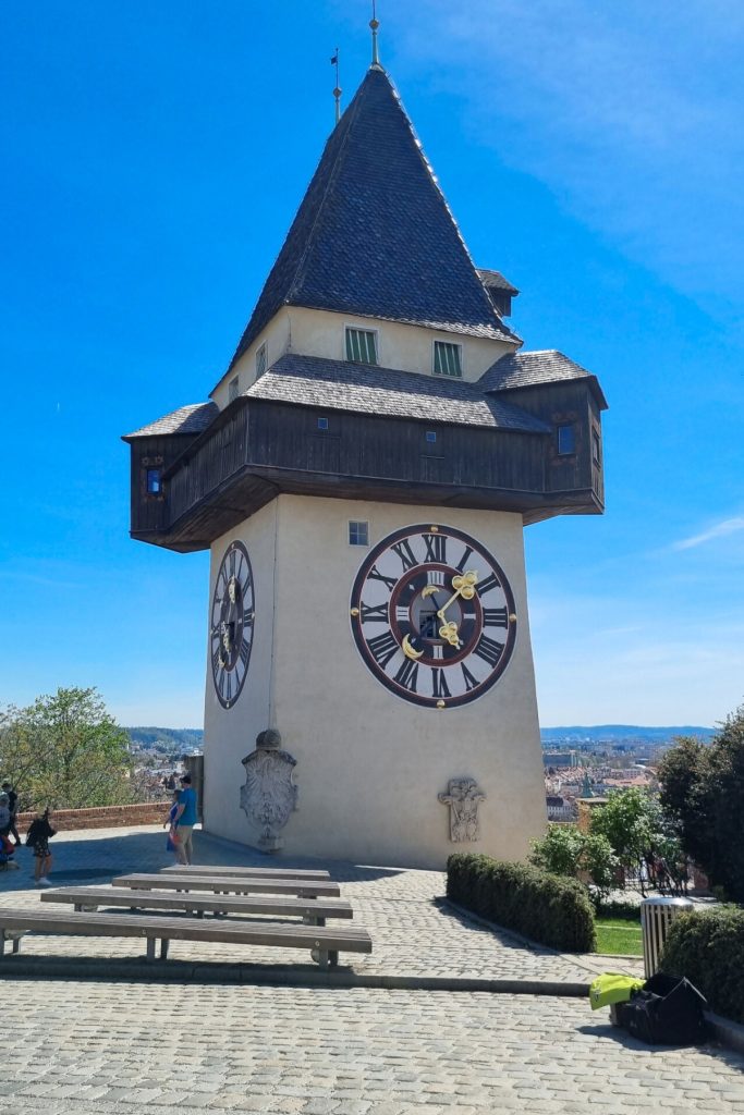 The Graz Clock Tower rises above a cobblestone terrace under a bright blue sky, with large clock faces visible on two sides and the city spread out in the distance below. A few visitors stand near the base, showing this as a hilltop landmark and viewpoint.