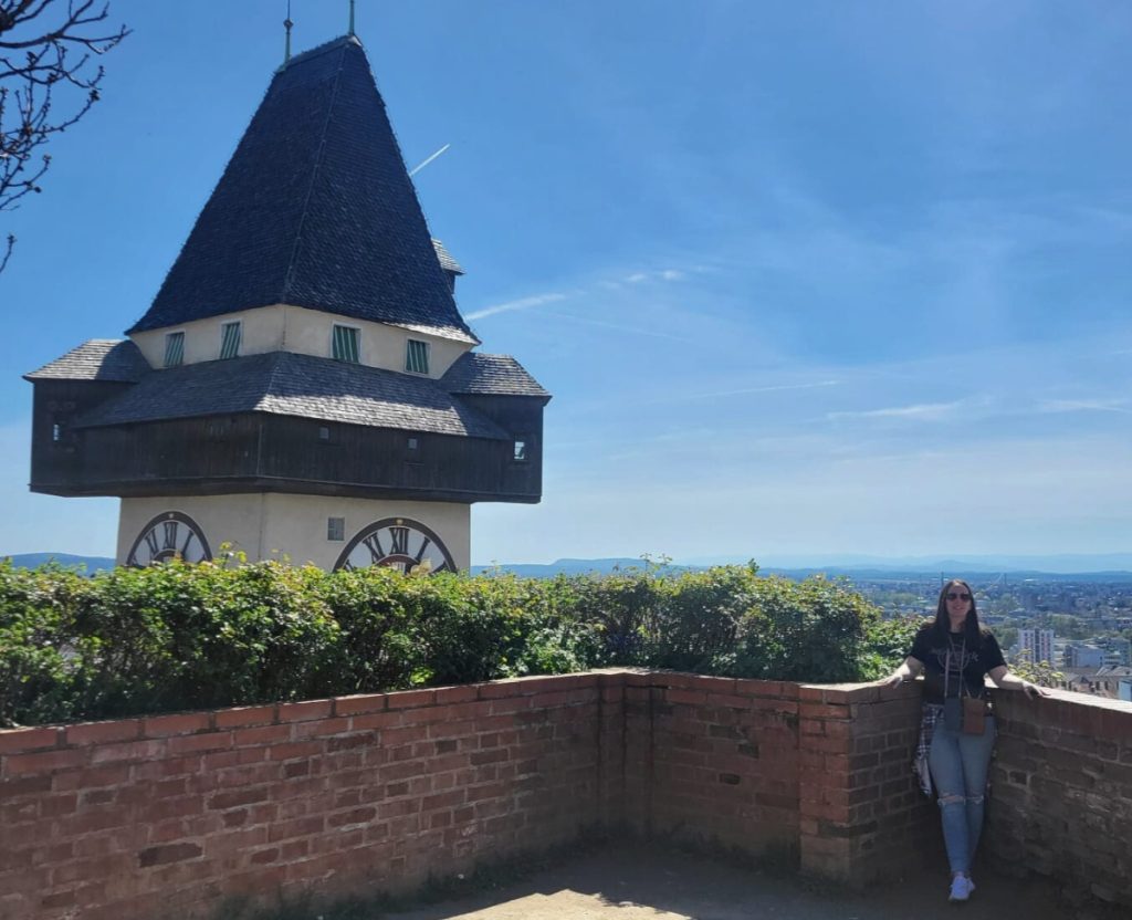 The Graz Clock Tower rising above a brick lookout terrace on Schlossberg hill with a panoramic city view in the background. A woman stands at the corner of the terrace enjoying the viewpoint.