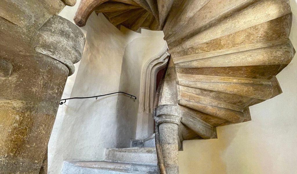 Stone spiral staircase curves tightly upward inside a historic tower, with thick columns, worn steps, and pale plaster walls enclosing the space. The view from below emphasizes the twisting shape and old architectural details of the stairwell.