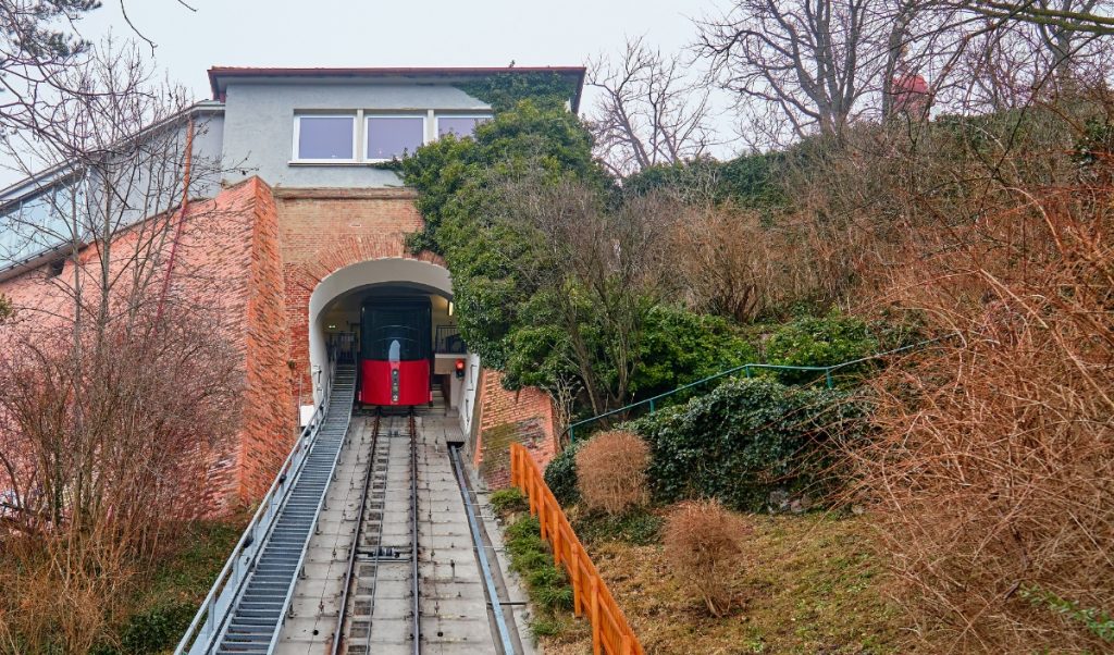 Red funicular car emerging from a brick tunnel on a steep hillside with tracks running down the center. Bare winter trees and thick shrubs surround the station, showing a hillside transit stop that could fit into a Graz itinerary.