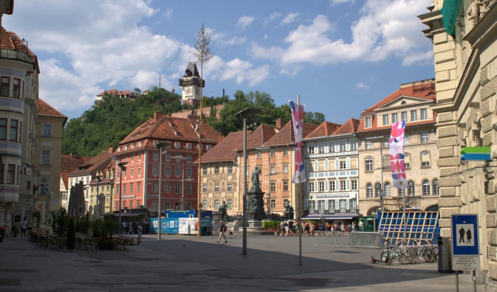 Wide view of Graz’s main square framed by pastel historic buildings and open paving, with the Clock Tower visible on the green hill in the background. People gather around the central monument while red tiled rooftops and old facades give the square a classic old town feel.
