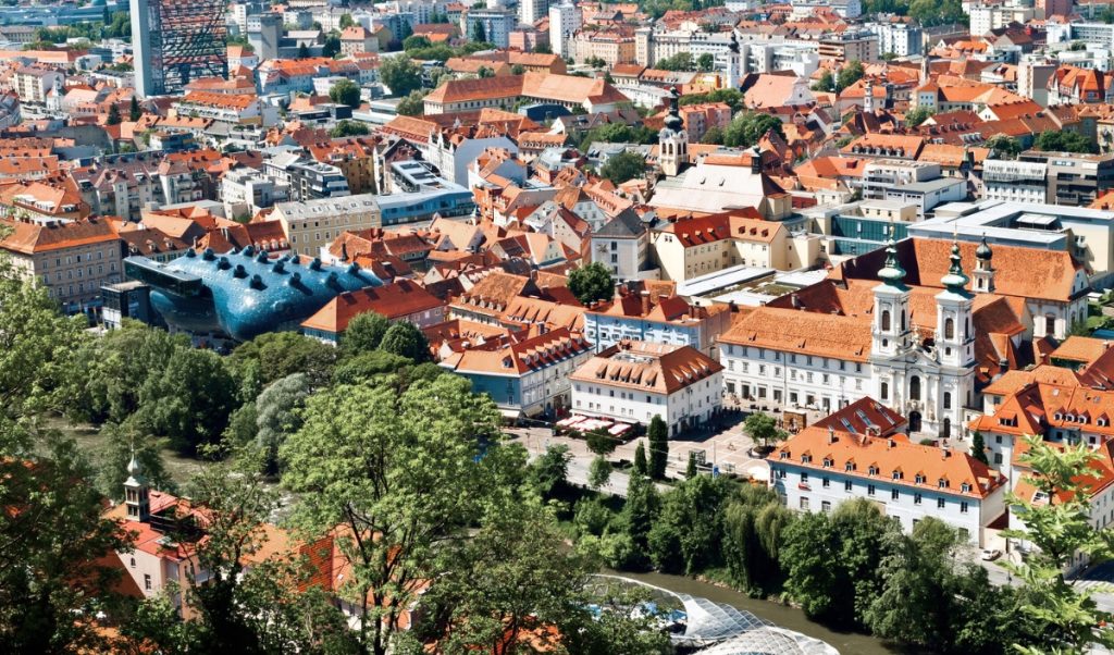 Panoramic view over Graz with dense orange tiled rooftops, church towers, and the Mur River lined with green trees in the foreground. The modern blue Kunsthaus stands out among the historic old town buildings, making this a strong overview scene for a Graz itinerary.