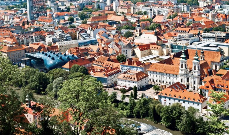 Panoramic view over Graz with dense orange tiled rooftops, church towers, and the Mur River lined with green trees in the foreground. The modern blue Kunsthaus stands out among the historic old town buildings, making this a strong overview scene for a Graz itinerary.
