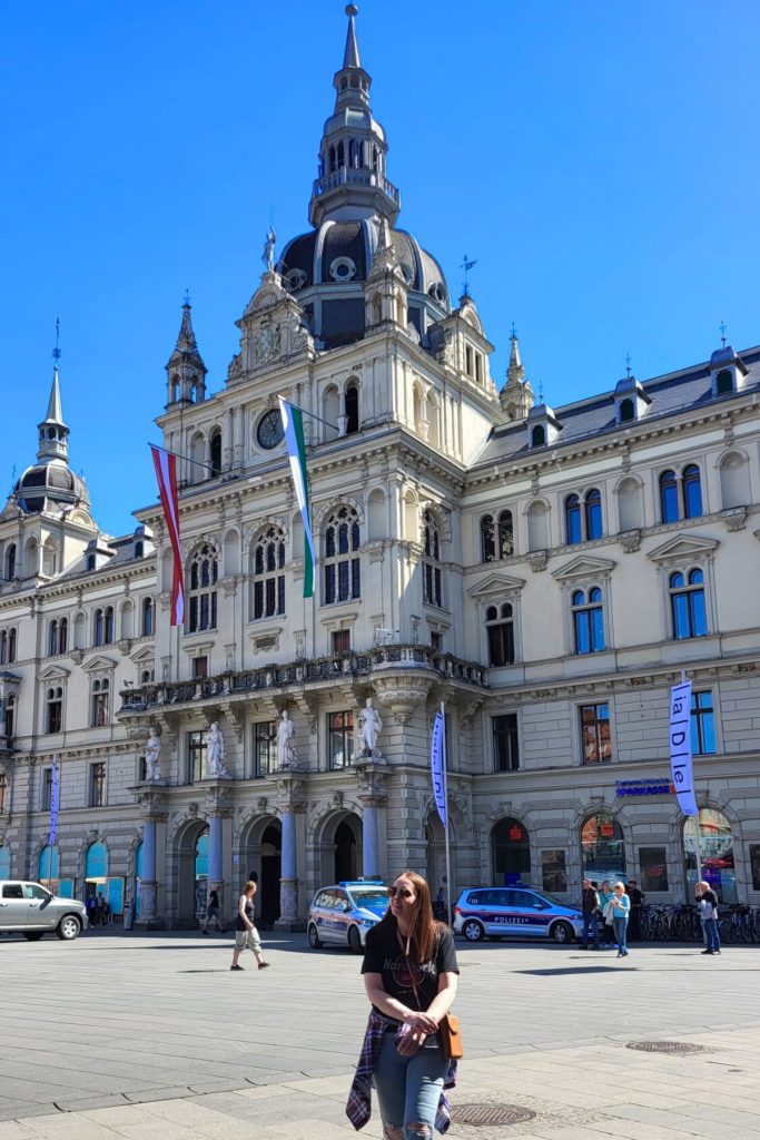 Grand historic city hall building with ornate façade and clock tower rising above the main square in Graz. Austrian flags hang from the balcony while a woman stands in the plaza below.