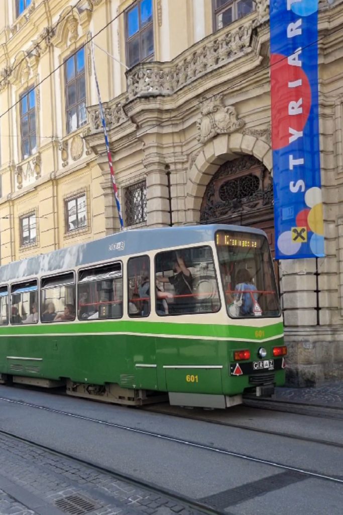Green and white tram driving along tracks past an ornate historic building in Graz. A tall blue banner on the building reads "STYRIA".