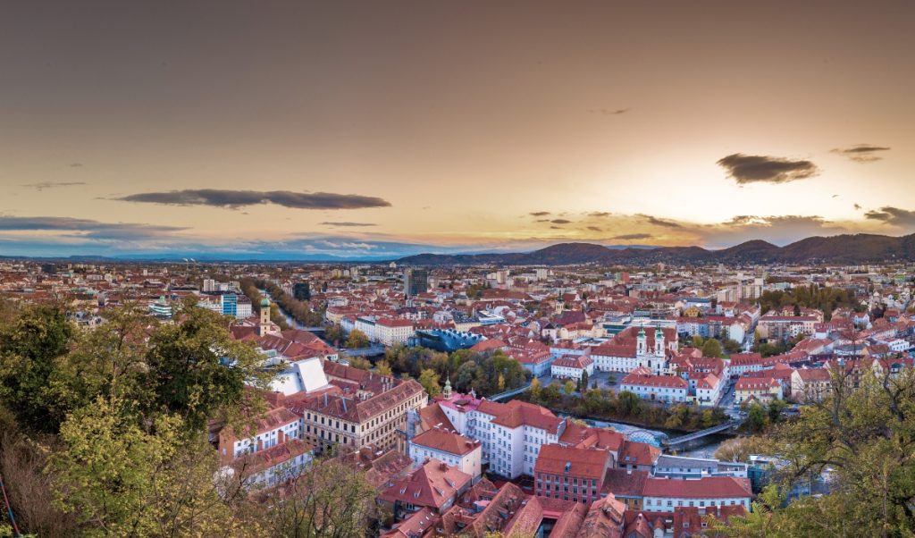 Panoramic view over Graz at sunset with red tiled rooftops spreading across the city and low hills on the horizon. Trees frame the foreground while warm evening light and a glowing sky make the skyline feel expansive and calm.