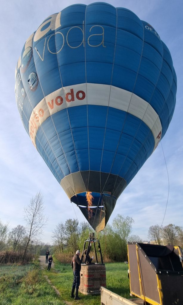 A blue hot air balloon floats just above a grassy field with a burst of flame visible inside the envelope above the basket. The balloon shows the text "balso vodo" on a white band while a man stands beside the wicker basket and a black trailer sits nearby.