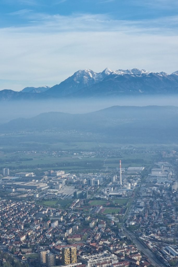 Snow covered mountains rise behind a hazy city with factories, apartment buildings, and rail lines in the foreground. The layered hills and mist make the mountain range stand out above the urban area.
