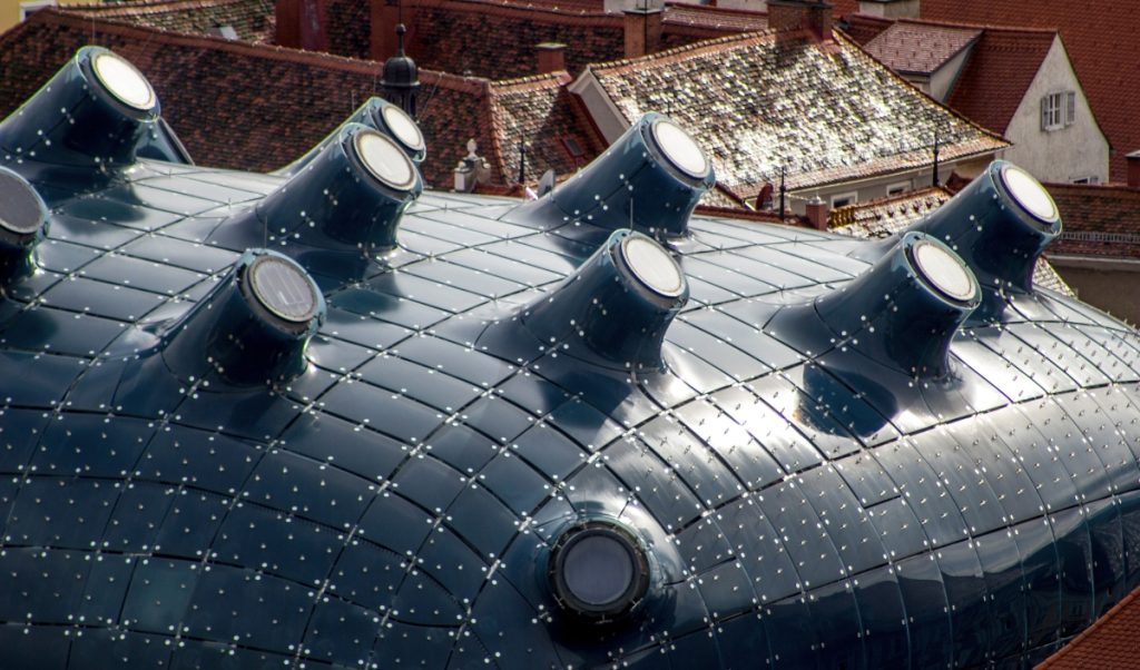 Close view of the Kunsthaus Graz roof with its glossy dark blue curved surface and round nozzle like skylights rising across the top. Red tiled historic rooftops in the background highlight the contrast between the museum’s futuristic design and the surrounding old town.
