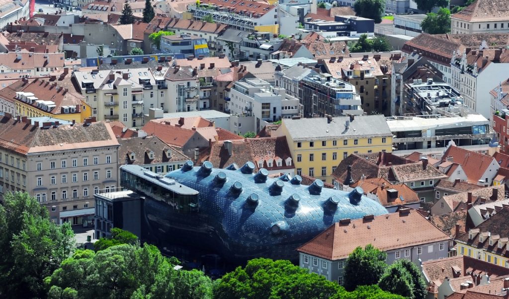 Elevated view of Graz with the dark blue Kunsthaus museum surrounded by tightly packed historic buildings and red tiled rooftops. Trees frame the lower edge of the scene, and the museum’s rounded futuristic shape stands out sharply against the old town around it.
