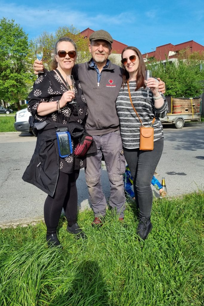 My sister and I with our pilot standing on a grassy roadside holding champagne flutes and smiling at the camera. The man in the middle wears a flat cap and dark jacket while the two women beside him wear sunglasses, with a parked trailer and buildings behind them.