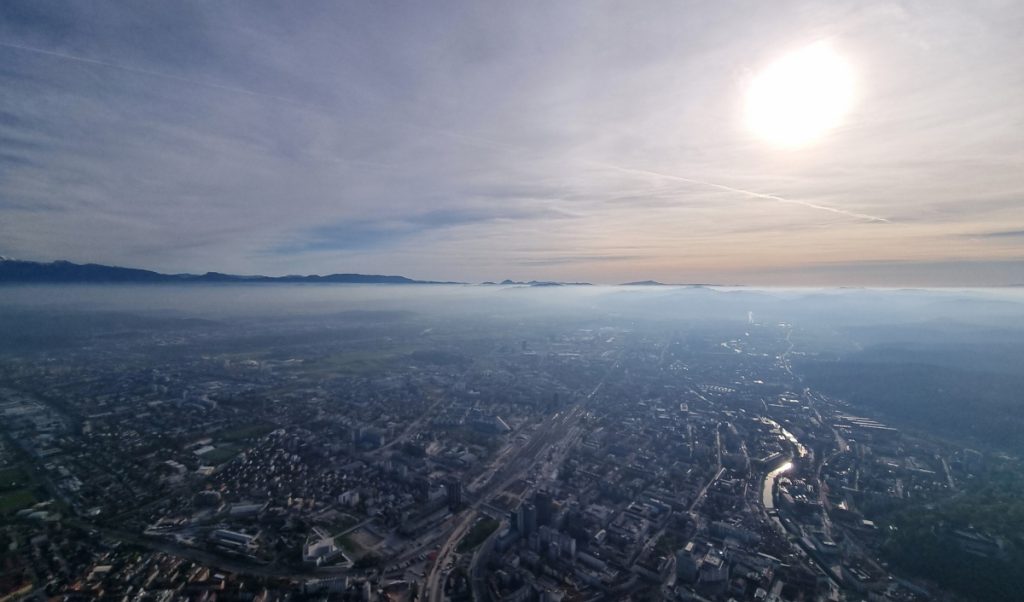 High aerial view over a hazy cityscape with the sun glowing through thin cloud and mist. A river reflects light as it winds through the darker buildings and streets below.