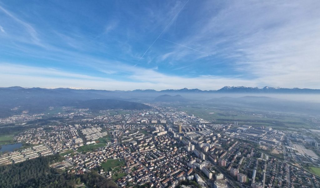 Wide aerial view of a dense city stretching across a valley with snow capped mountains lining the horizon. Wispy clouds streak the sky above neighborhoods, roads, and industrial buildings below.