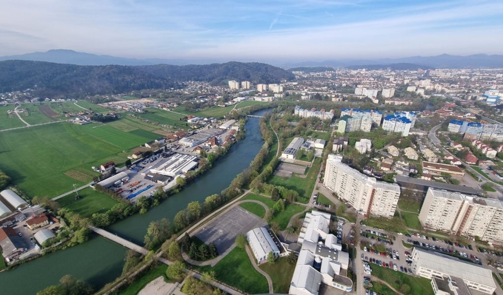 Aerial view of a city neighborhood divided by a green river with bridges, apartment blocks, warehouses, and fields spreading toward wooded hills. The scene shows the mix of urban streets and open farmland from high above.