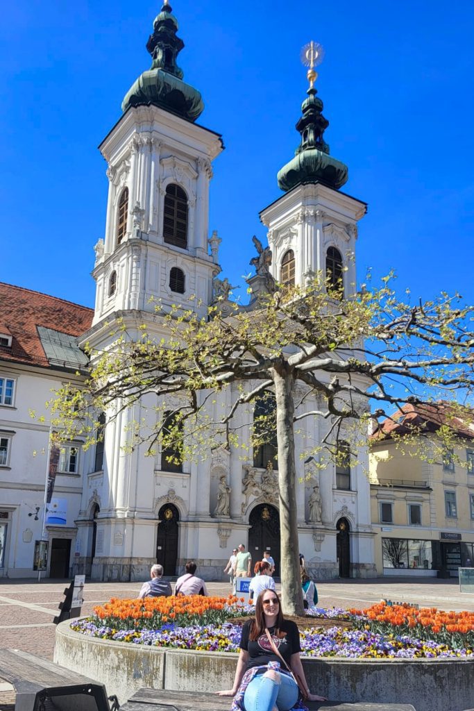 Woman sitting beside a circular flower bed of orange tulips in front of a white baroque church with two towers in Graz. People walk across the open square under a clear blue sky.