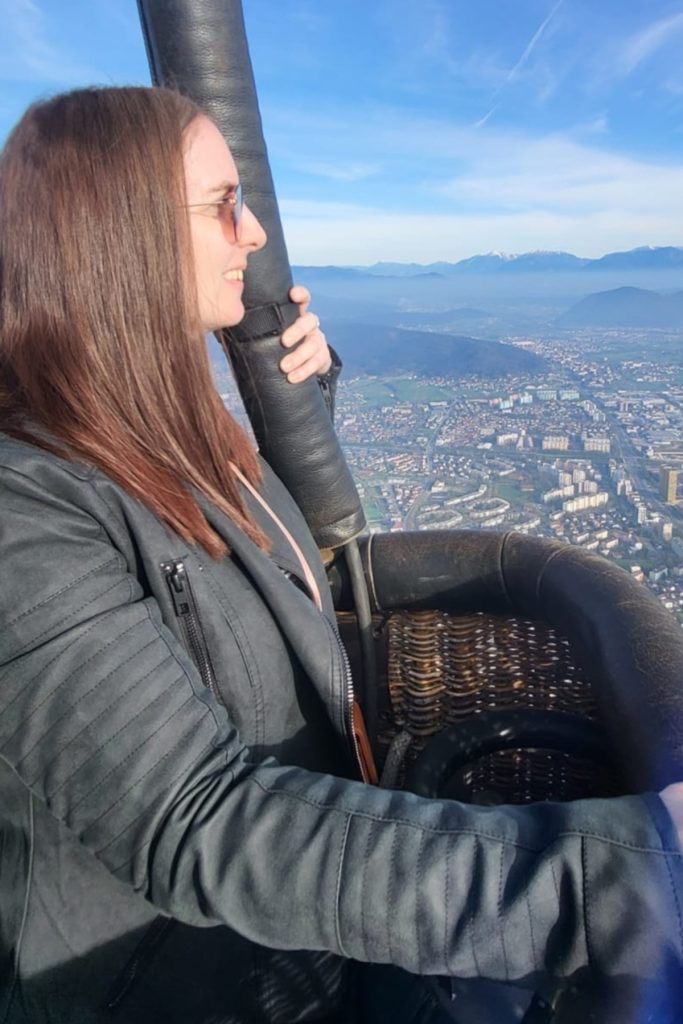 A woman wearing sunglasses smiles from the basket of a hot air balloon while looking out over a city spread across the valley below. Blue mountains and a pale sky frame the view behind her.