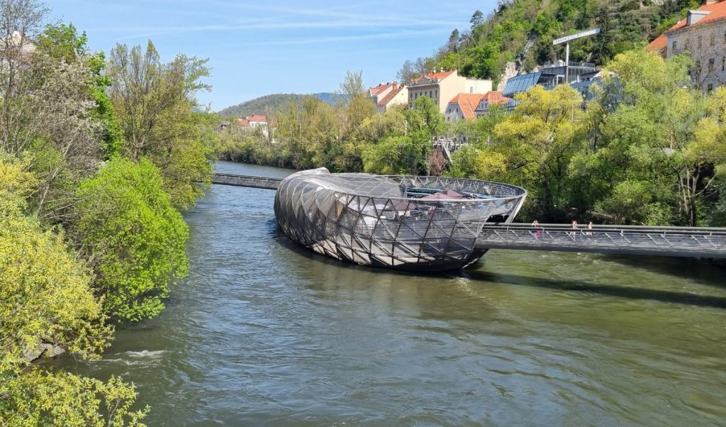 Futuristic steel island structure with a curved lattice shell floating in the Mur River in Graz. A pedestrian bridge connects the island café to the riverbank lined with green trees.