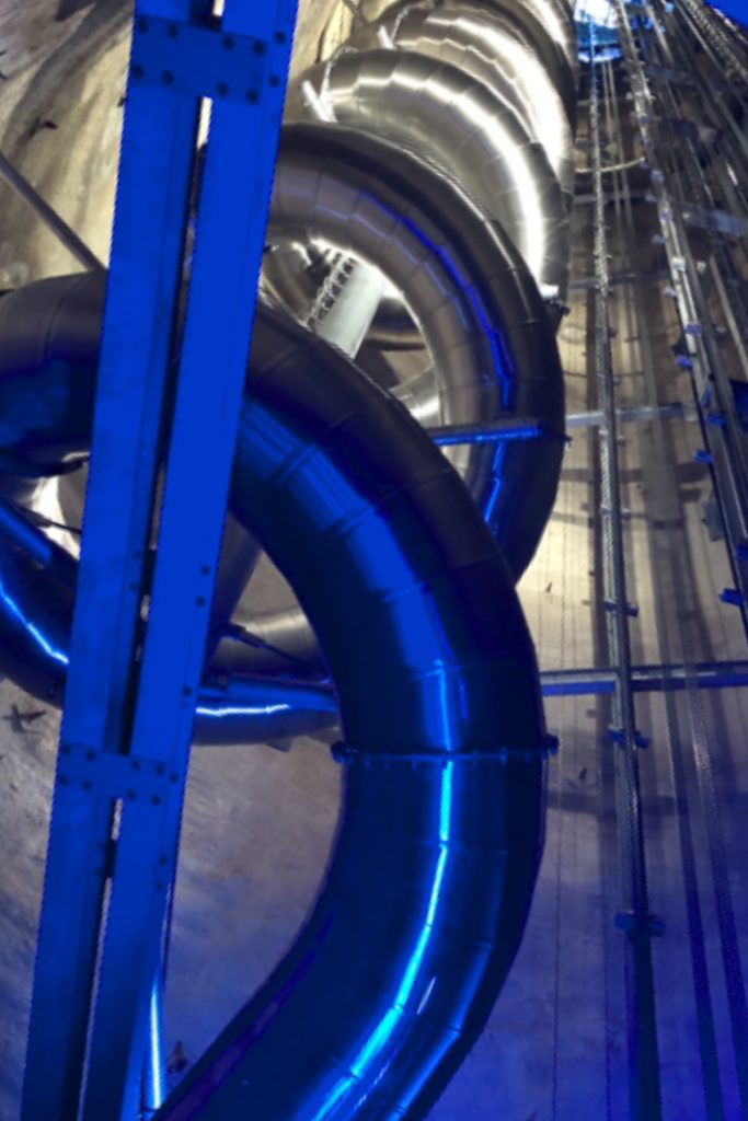 A shiny metal spiral slide winds downward inside a tall concrete shaft lit with blue and white lights. The steep upward angle emphasizes the height of the structure and the dramatic indoor design.