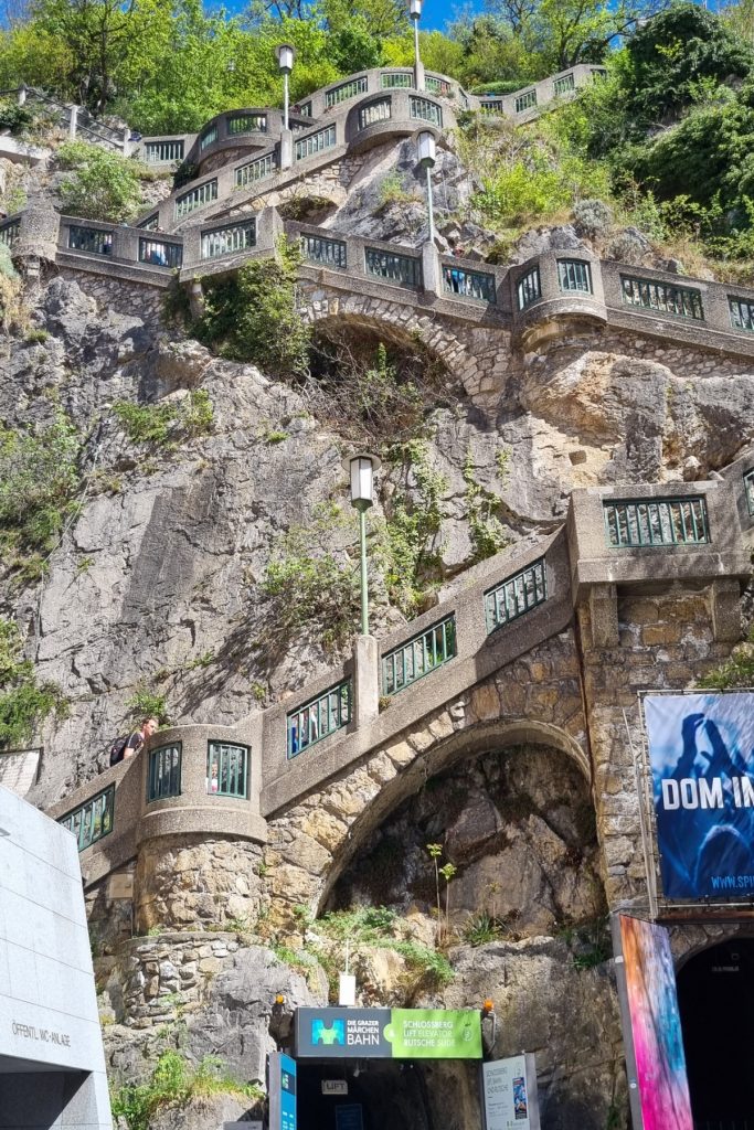 A steep stone staircase zigzags up a rocky hillside with terraces, arches, and railings built into the cliff. At the bottom, signs mark the Schlossberg lift and slide entrance, showing the dramatic uphill path to one of Graz’s hilltop attractions.