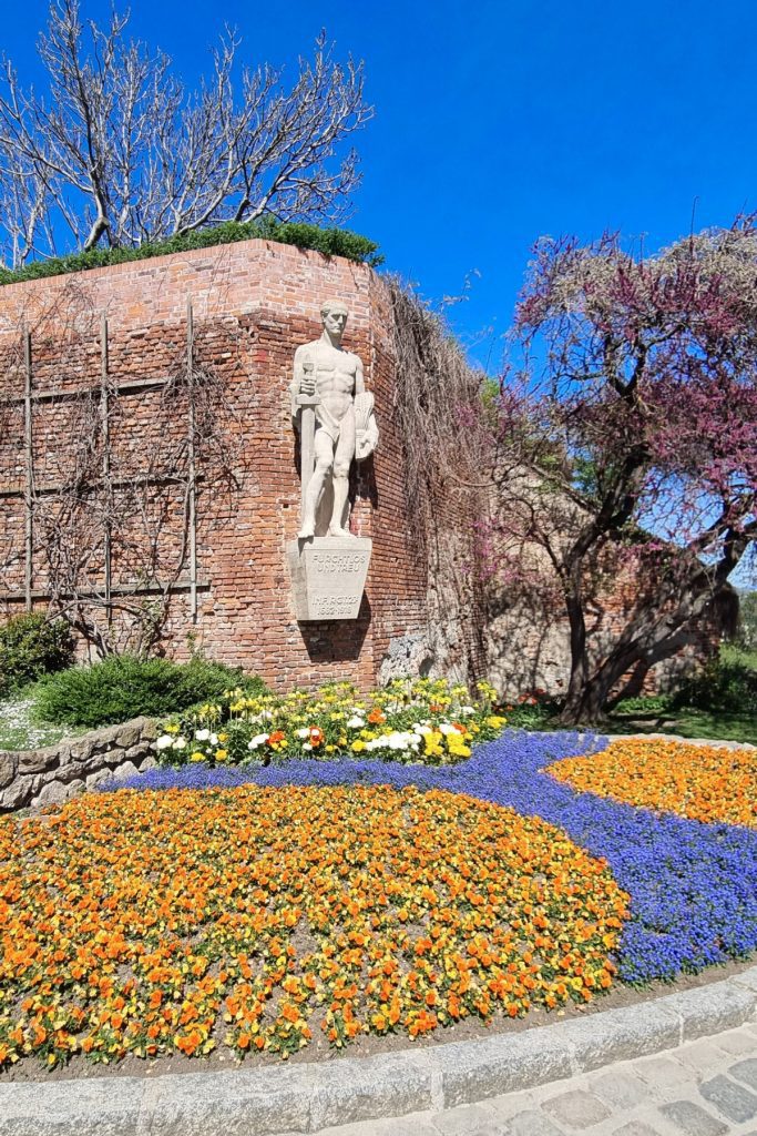 Stone statue mounted on a brick wall above colorful flower beds of orange, yellow, and purple in a landscaped garden on Graz Schlossberg. Trees and blue sky frame the historic hilltop park.