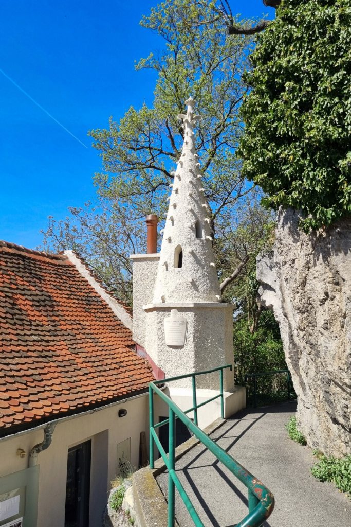 Narrow paved path with green railing leading past a small white tower and red tiled roof on the hill near Graz Clock Tower. Trees and bright blue sky surround the hillside viewpoint.