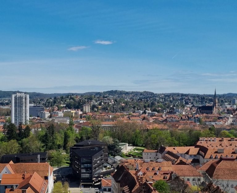 Wide panoramic view of Graz showing red tiled rooftops, church spires, and modern buildings surrounded by green hills. The city stretches across the valley under a clear blue sky.