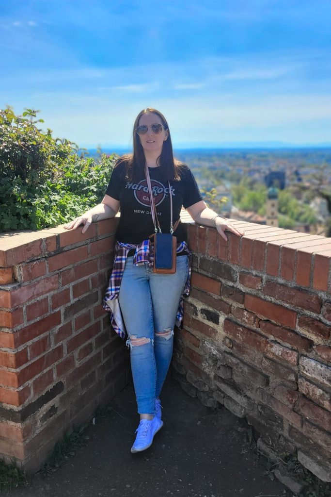 This is me wearing a shirt that says "Hard Rock Cafe New Delhi" stands at a brick lookout wall on Schlossberg hill above Graz. The city spreads out behind her under a bright blue sky.