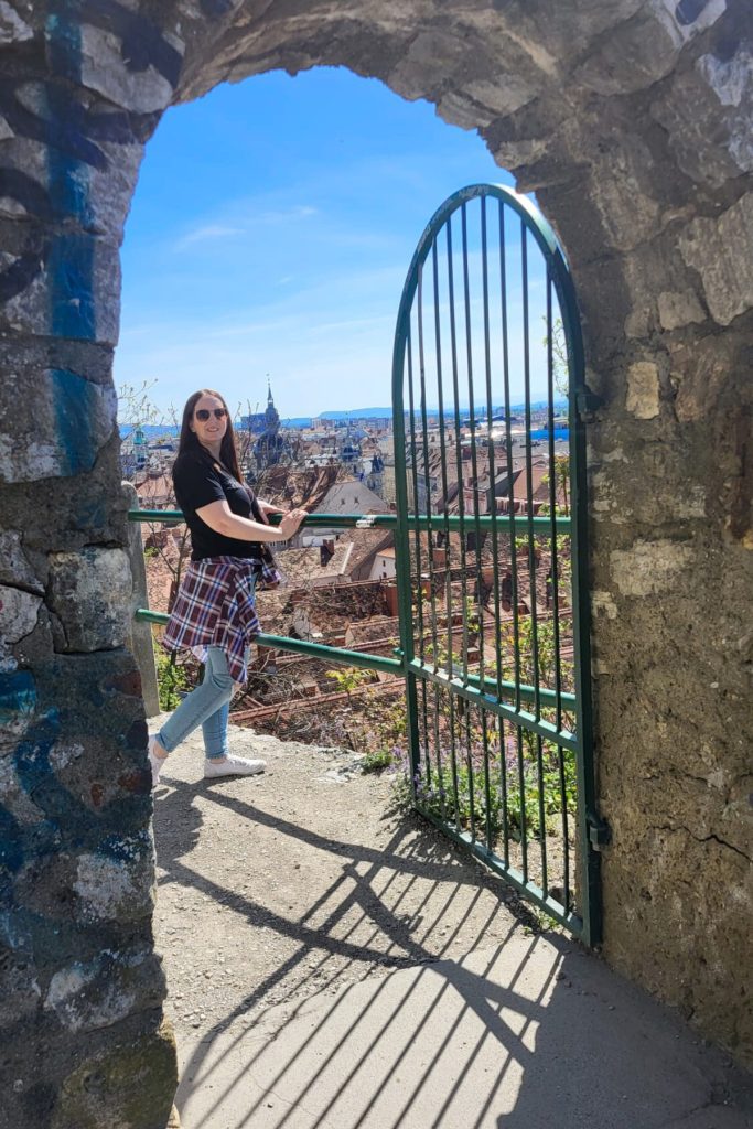 Woman standing beside an iron gate framed by a stone arch overlooking the red rooftops of Graz. Church spires and historic buildings stretch across the city skyline in the distance.