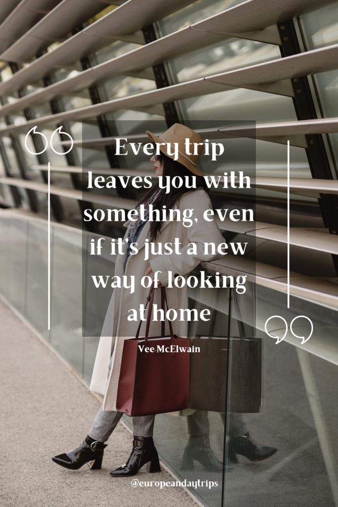 Traveler in a tan hat and cream coat leans against a glass wall with metal slats, carrying a burgundy tote bag, with her reflection visible beside black ankle boots and plaid trousers. Overlaid text reads "Every trip leaves you with something, even if it’s just a new way of looking at home" and "Vee McElwain" with "@europeandaytrips" at the bottom.
