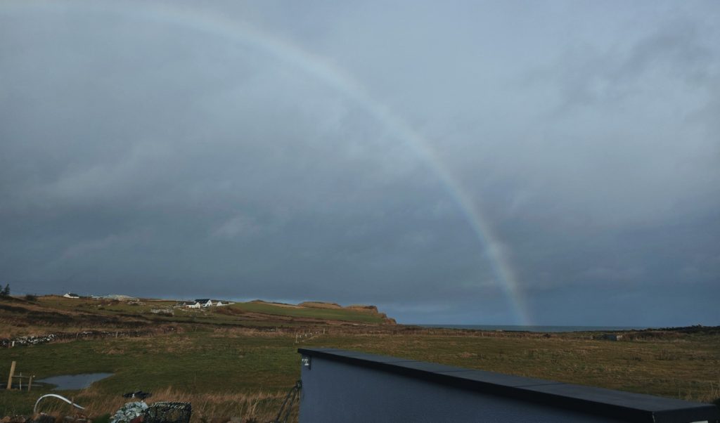 Faint rainbow arcs across a dark cloudy sky over open fields and low cliffs in rural Ireland. This moody landscape reflects the changeable weather to expect when planning what to pack for a trip to Ireland.