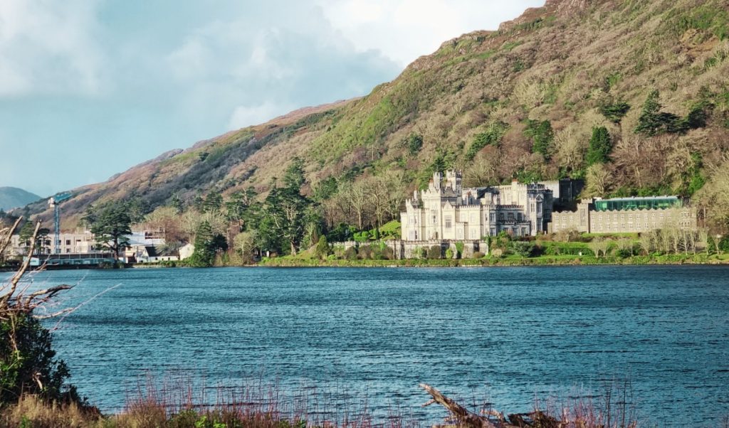 Lakeside view in Ireland with a historic stone castle at the base of a green hillside across bright blue water. The sunny scene highlights a classic stop on an Ireland trip and the kind of layered weather conditions to keep in mind when packing.
