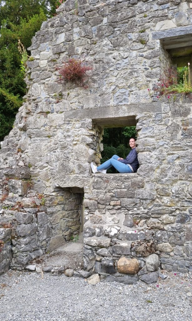 Me sitting in a stone window opening inside old castle ruins in Ireland, surrounded by rough gray masonry and a gravel courtyard below. Her sweater, jeans, and white sneakers show a casual outfit that works well for how to pack for a trip to Ireland when visiting historic sites.