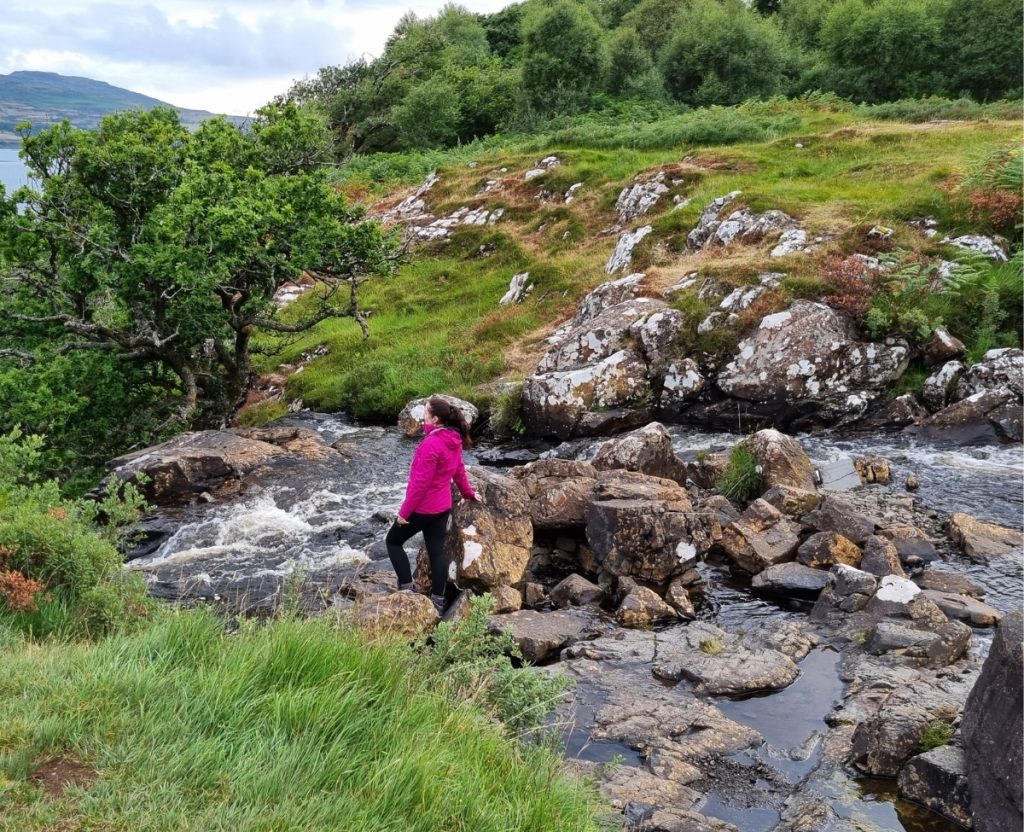 Me in a bright pink rain jacket stands on rocks beside a rushing stream in the Irish countryside, surrounded by green hills, trees, and uneven terrain. The scene shows why sturdy shoes and a waterproof layer matter when deciding how to pack for a trip to Ireland.