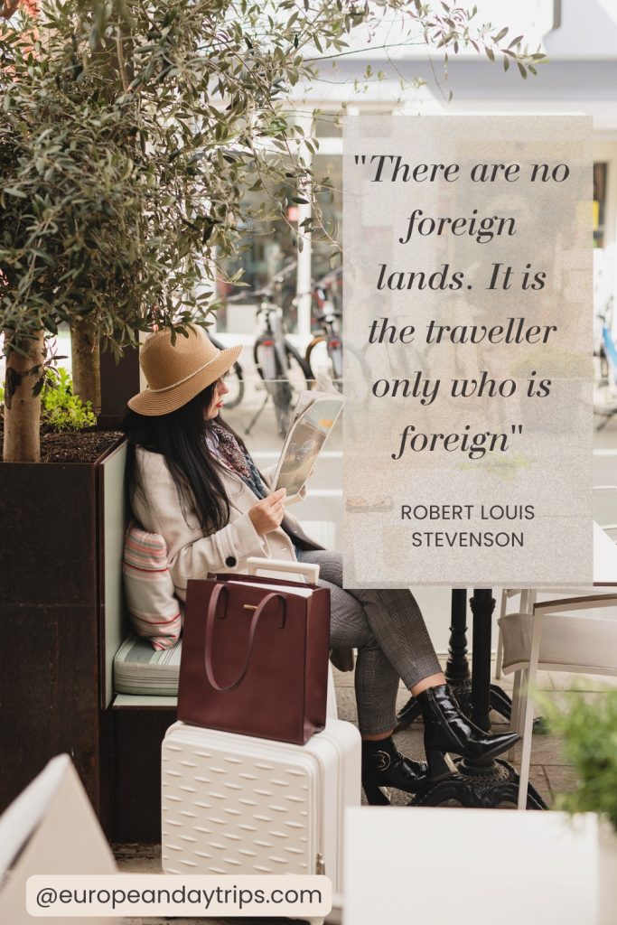Traveler in a tan hat and light coat sits at an outdoor café bench reading, with a white suitcase and brown tote bag beside her under leafy trees. Overlaid text reads "There are no foreign lands. It is the traveller only who is foreign" and "ROBERT LOUIS STEVENSON" with "@europeandaytrips.com" at the bottom.