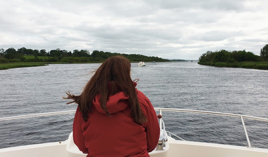 Traveler in a red rain jacket stands at the front of a boat on a wide river in Ireland beneath a gray overcast sky. The scene shows the kind of cool wet weather to pack for on an Ireland trip, with tree lined banks and a few small boats ahead.