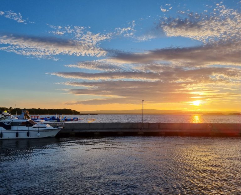 Sunset over a marina in Ireland with two white boats moored beside a long dock and calm water reflecting the orange light. Blue sky and scattered clouds stretch across the scene while the sun sits low on the horizon, capturing a peaceful moment on an Ireland trip.