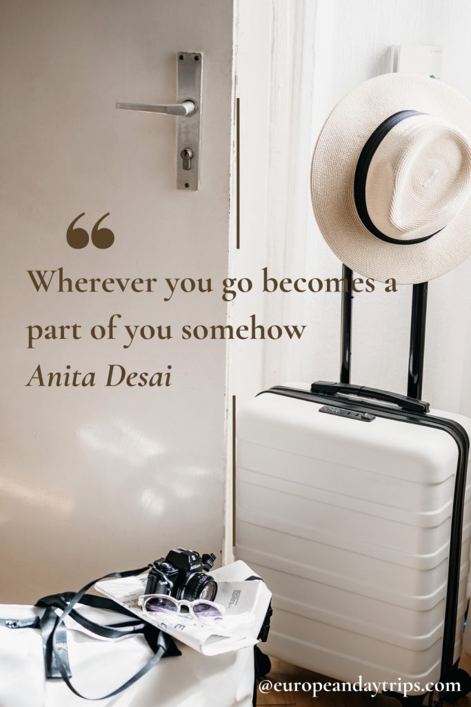 Open white door beside a white hard shell suitcase with an extended handle and a straw hat resting on top, with a camera, sunglasses, and papers placed on a white bench in the foreground. Overlaid text reads "Wherever you go becomes a part of you somehow" and "Anita Desai" with "@europeandaytrips.com" in the bottom corner