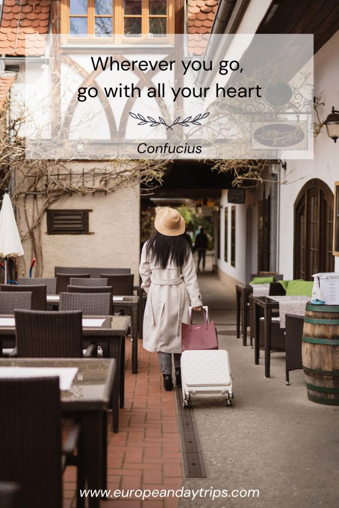 Narrow old town walkway lined with outdoor café tables and chairs, where a traveler in a beige coat and tan hat pulls a white suitcase and carries a pink bag beneath timbered buildings. Overlaid text reads "Wherever you go, go with all your heart" and "Confucius" with europeandaytrips.com at the bottom.