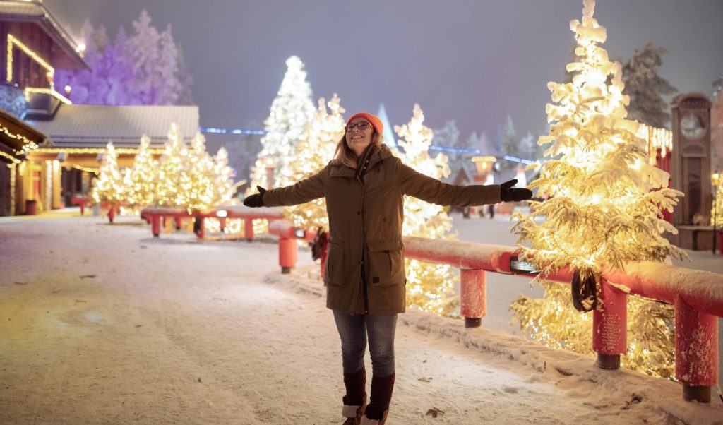 A woman stands with her arms out in a snowy Christmas village filled with glowing trees and festive lights.