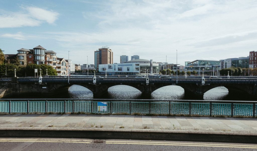 A wide bridge crosses a calm river with city buildings and apartments in the background. A teal railing and quiet street frame the waterfront view.