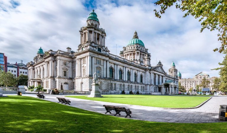 Belfast City Hall with green copper domes behind a wide lawn and benches on a sunny day. A classic stop on a day trip from Dublin to Belfast.