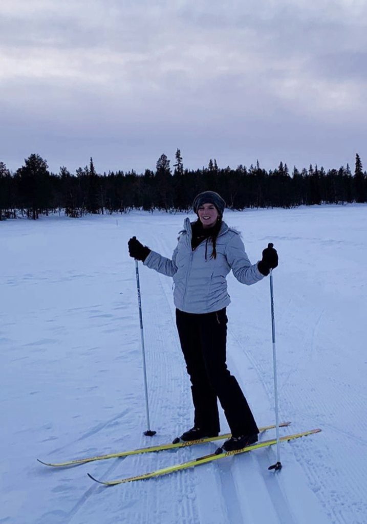 A woman smiles while cross country skiing across a snowy open trail, with a dark pine forest in the distance.