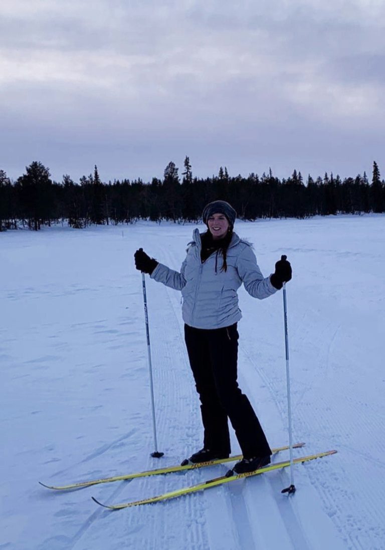A woman smiles while cross country skiing across a snowy open trail, with a dark pine forest in the distance.