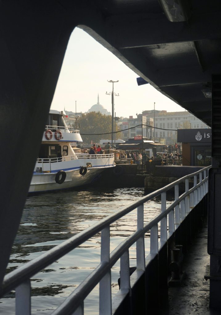 Ferry dock in Istanbul viewed from beneath a shaded pier, with railings leading toward the water and a white ferry tied up nearby. A mosque dome and minaret appear hazy in the distance behind the busy waterfront.